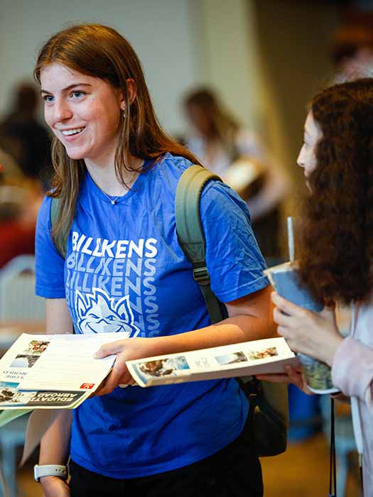 A smiling Billiken with long hair holds an admission booklet