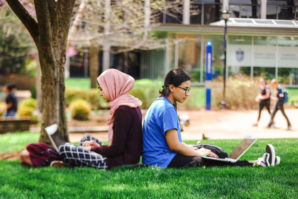 two students, one wearing a headscarf, sit back to back in the grass under a tree