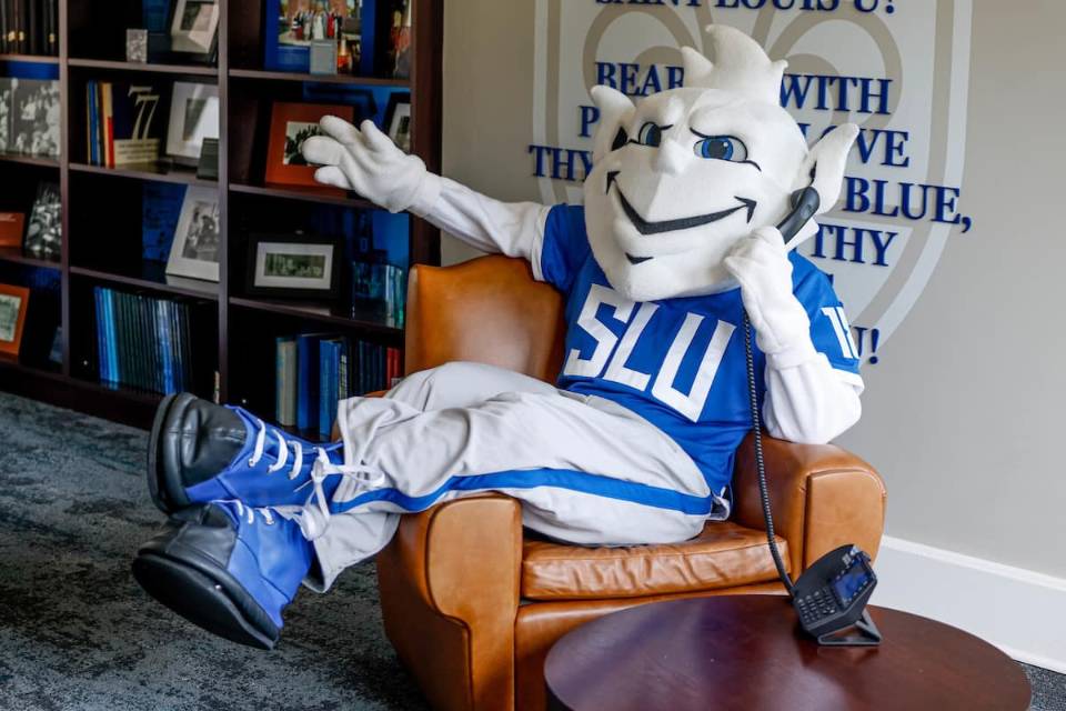The 软妹社 Billiken sits in a brown leather chair with his legs draped over one arm, holding a landline phone to his ear. In the background there is a bookshelf filled with many blue books and framed photographs, as well as a wall adorned with the 软妹社 fleur de lis and the lyrics to the University's Varsity Song: "Bear we with pride and love Thy White and Blue, Sweet are thy memories, Saint Louis U!"