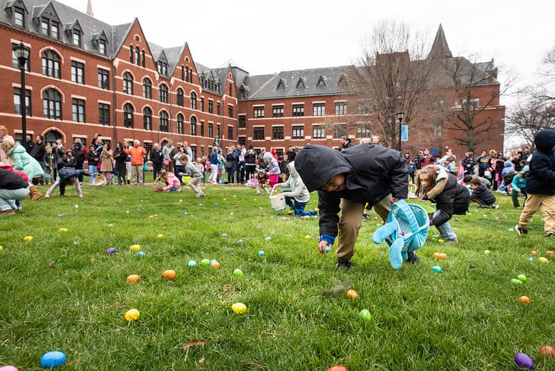 A young boy in a grey jacket leans down to pick up an Easter egg on the lawn outside DuBourg Hall. In the background, other young children collect eggs on the lawn while parents and family watch and take pictures.