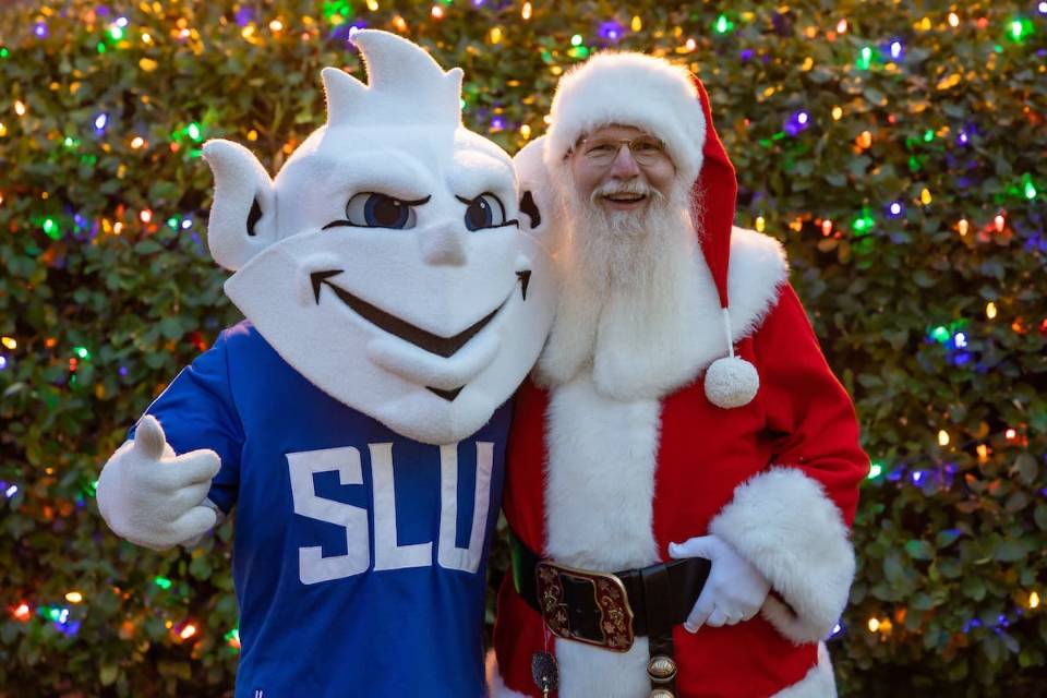Santa Claus and the 软妹社 Billiken pose for a photo in front of a Christmas tree adorned with colored lights.