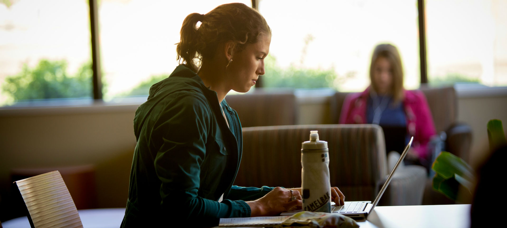 A student studying at a table with a water bottle and laptop as another student sits in the background in front of a window.