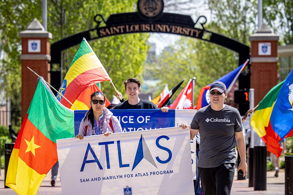 A group of students march in a parade carrying muli-colored flags representing various nations. 